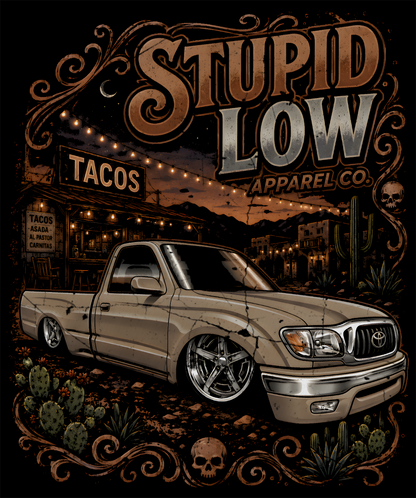 A stylized graphic print featuring a lowered beige pickup truck in a desert town scene with large text reading 'STUPID LOW APPAREL CO.' The artwork includes decorative swirls, cacti, skulls, and warm string lights against a dark background.